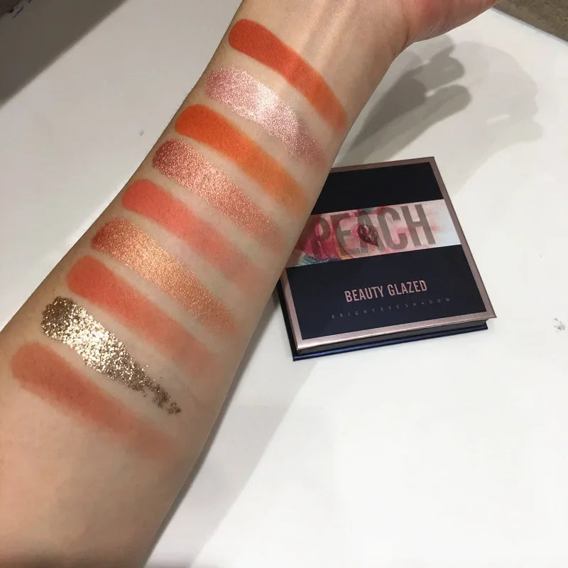 Nine-color eyeshadow palette with shimmer and matte shades swatched on a forearm, featuring warm orange and gold tones.